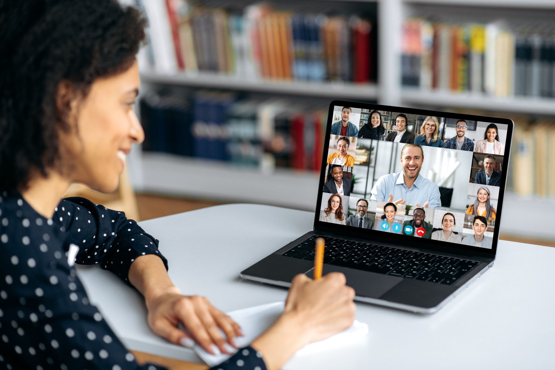 Video call, online conference using app and laptop. African American female student, studying remotely, watches an online lecture, taking notes, multiracial smiling people on laptop screen